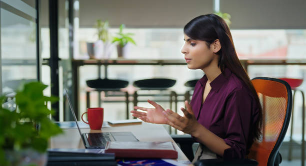 Indian young adult gen z busy business woman lady sitting on chair use laptop look screen do online work video call talk indoor office place. Teen expert female staff worker distant conference conversation project discussion meeting centre job workplace